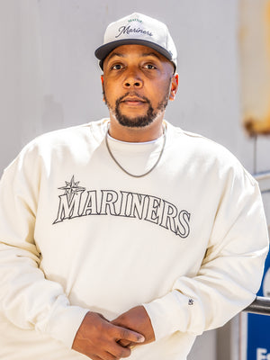 Man wearing a Mariners cream hoodie and  Mariners golfer hat.